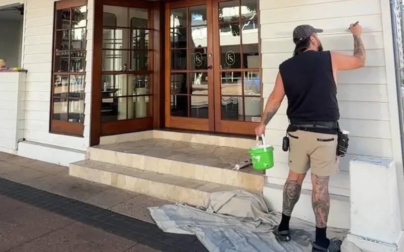 Aaron brush painting weatherboards on a shopfront, with drop sheets down