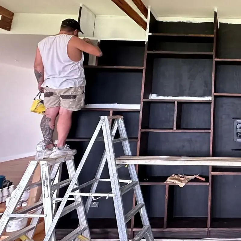 Aaron working from ladders and a plank to paint a full-height built-in bookshelf